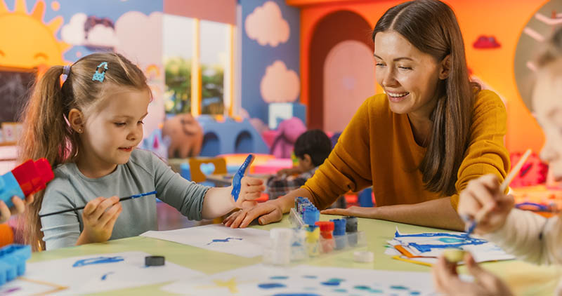 Portrait Of Adorable Girl Using Watercolor To Create Colorful Paintings On Paper. Cheerful Female Teacher Spending Productive Time In Preschool, Teaching Kids To Paint In Art Class